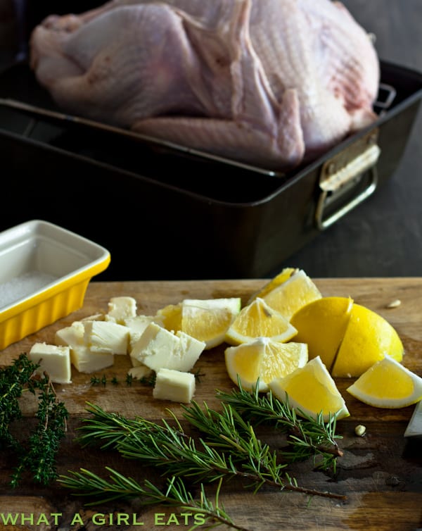Raw turkey in a pan with lemons, herbs, and butter on a cutting board nearby.