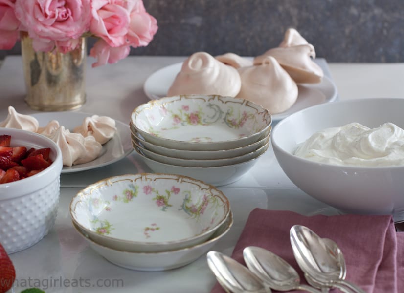 Pretty china bowls and spoons on a table with meringues, cream and strawberries.