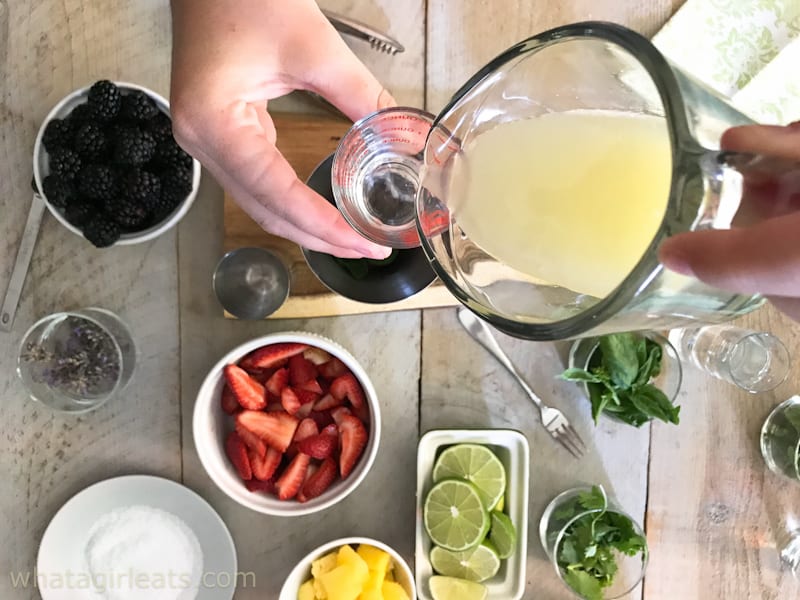 Pitcher of margaritas being poured into a cocktail shaker, with fruits and herbs on a table.