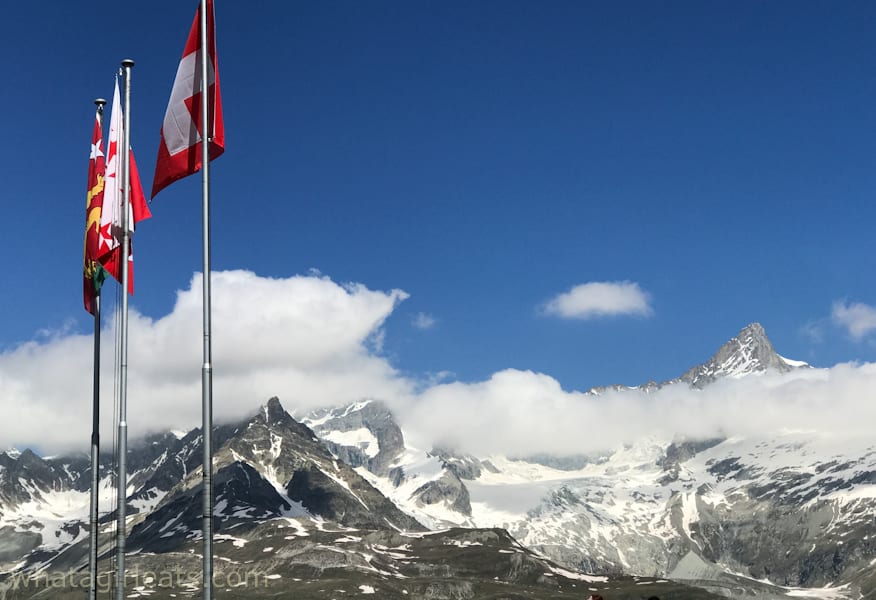 Flagpoles with snow-covered mountains in the background and clouds rolling in.