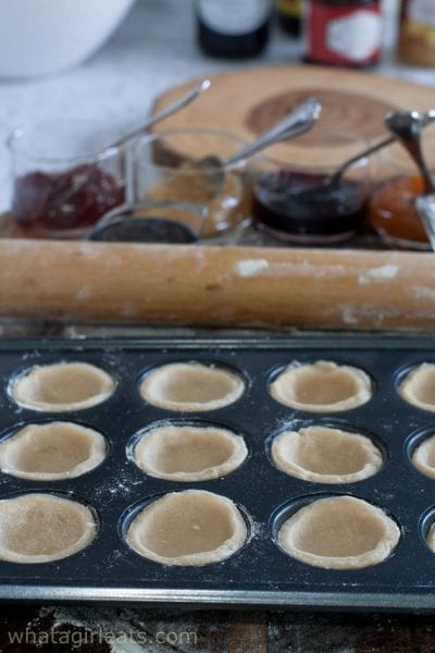 Small bowls of jam, dough pressed into mini muffin tins, and a rolling pin.