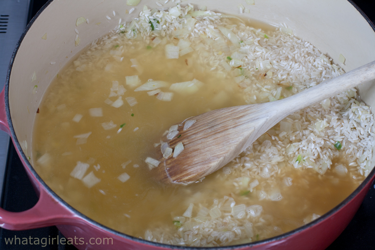 Rice, broth, and onions in a pot with wooden spoon stirring. 